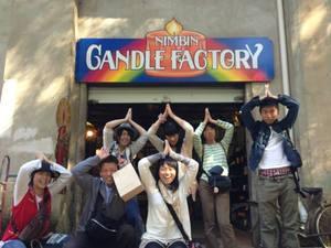 「CANDLE FACTORY」と書かれた看板が掲げられた入り口前で、派遣学生7人が頭の上で両手を合わせキャンドルの炎のようなポーズをとって記念撮影をしている写真
